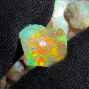 May include: A close-up of a rough opal gemstone with vibrant colors. The opal displays flashes of green, orange, and yellow, set against a milky white base. The stone is held in a black fabric, highlighting its natural beauty.