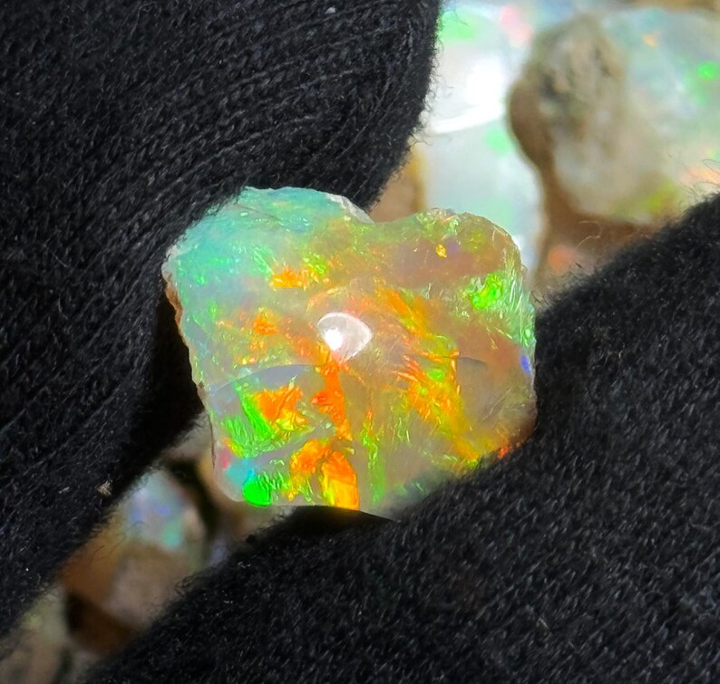 May include: A close-up of a rough opal gemstone with vibrant colors. The opal displays flashes of green, orange, and yellow, set against a milky white base. The stone is held in a black fabric, highlighting its natural beauty.