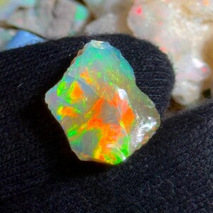May include: A rough-cut opal gemstone with vibrant colors of green, orange, and yellow. The gemstone is held against a dark background, showcasing its iridescent play-of-color. The opal's surface is uneven, revealing its natural, raw form.