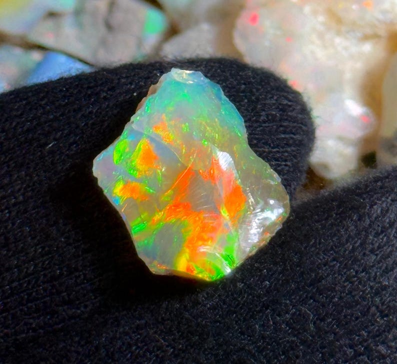 May include: A rough-cut opal gemstone with vibrant colors of green, orange, and yellow. The gemstone is held against a dark background, showcasing its iridescent play-of-color. The opal's surface is uneven, revealing its natural, raw form.