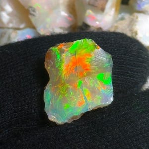 May include: A rough opal gemstone with vibrant colors. The opal displays flashes of green, orange, and yellow hues. The stone has a translucent appearance and a rough, natural texture. The opal is set against a dark background.