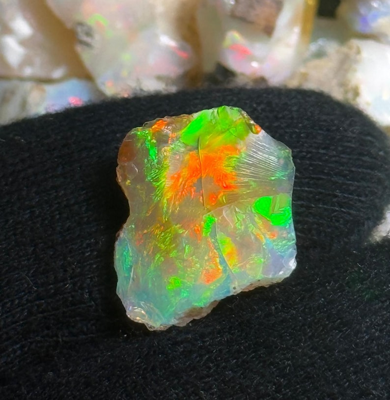 May include: A rough opal gemstone with vibrant colors. The opal displays flashes of green, orange, and yellow hues. The stone has a translucent appearance and a rough, natural texture. The opal is set against a dark background.