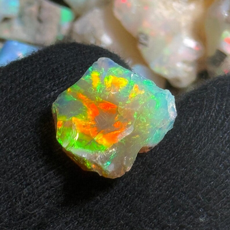 May include: A vibrant opal gemstone with a mix of colors, including green, orange, and yellow. The rough stone has a natural, irregular shape and is set against a dark background. The opal displays a play of color, reflecting light.