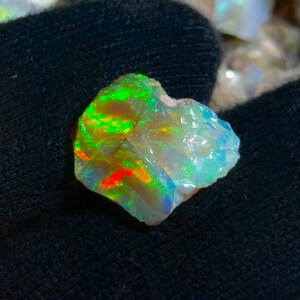 May include: A rough opal gemstone with vibrant colors. The stone displays flashes of green, red, and blue within its translucent, crystalline structure. The opal is set against a dark background, highlighting its iridescent qualities.