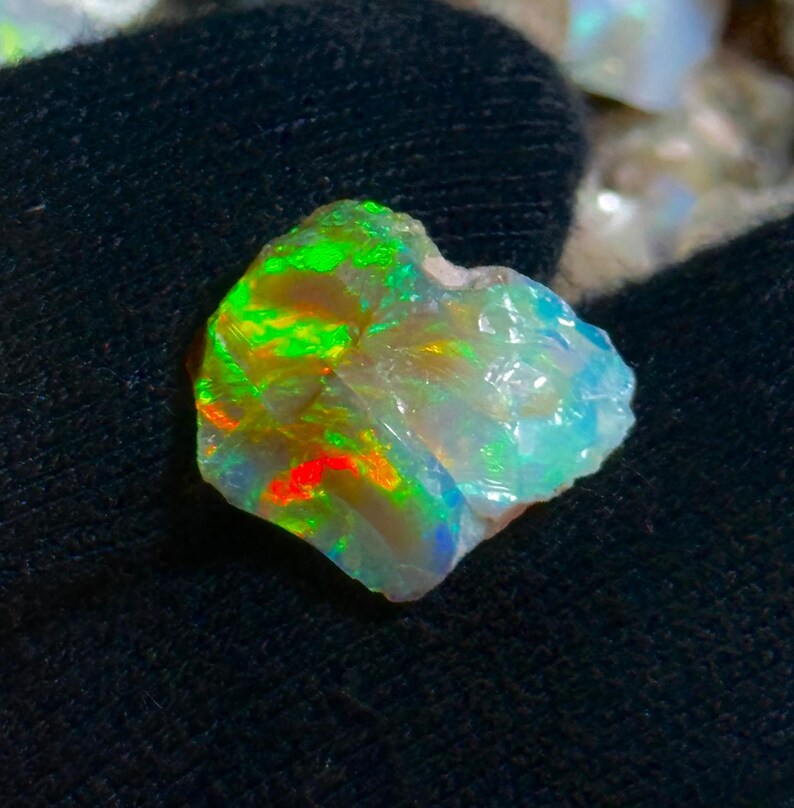 May include: A rough opal gemstone with vibrant colors. The stone displays flashes of green, red, and blue within its translucent, crystalline structure. The opal is set against a dark background, highlighting its iridescent qualities.