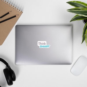 May include: A laptop with a sticker that says "Think SMART" in white and light blue. The laptop is on a white surface, with a notebook, pencils, headphones, a mouse, and a plant in the background.