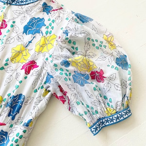 Puede incluir: Un vestido blanco con estampado floral con rosas azules, amarillas y rosas. El vestido tiene un ribete azul y blanco alrededor del cuello y las mangas.