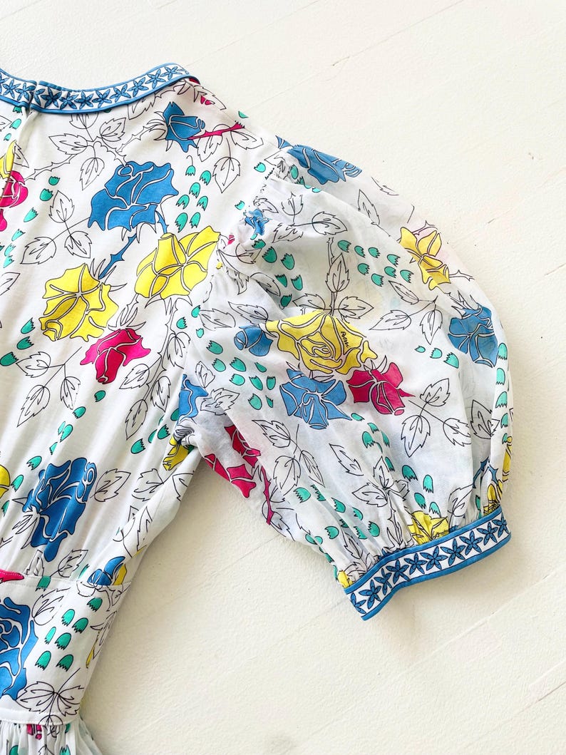 Puede incluir: Un vestido blanco con estampado floral con rosas azules, amarillas y rosas. El vestido tiene un ribete azul y blanco alrededor del cuello y las mangas.