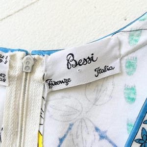 Puede incluir: Un vestido blanco con estampado floral azul y ribete azul. El vestido tiene una cremallera y una etiqueta que dice "Bessi Firenze Italia".