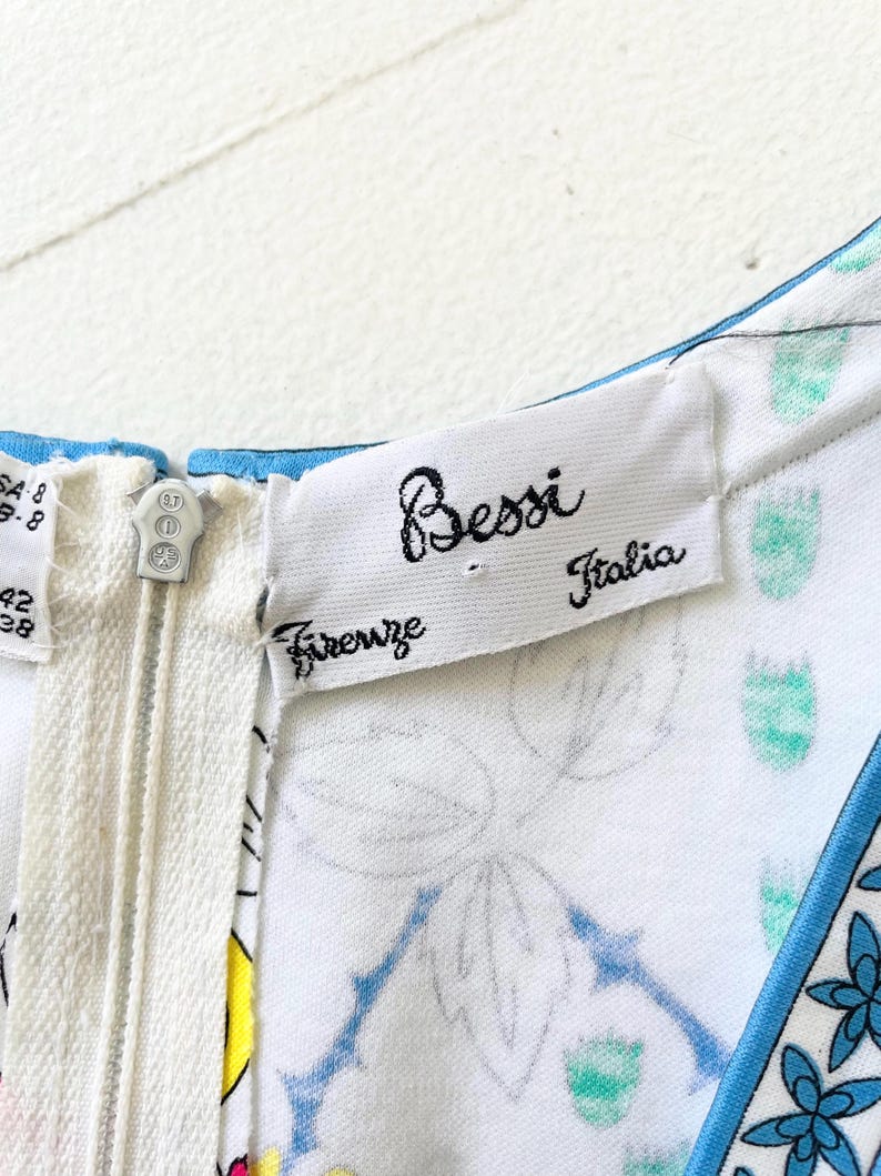 Puede incluir: Un vestido blanco con estampado floral azul y ribete azul. El vestido tiene una cremallera y una etiqueta que dice "Bessi Firenze Italia".