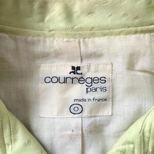 Puede incluir: Primer plano de una chaqueta verde claro con superficie texturizada. La chaqueta presenta una etiqueta que dice "courreges paris made in France". La chaqueta tiene cierre de bot&oacute;n.