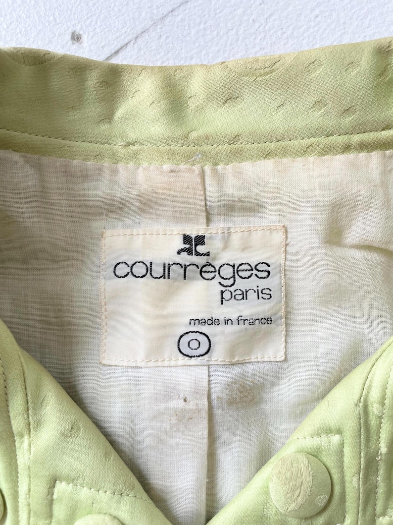Puede incluir: Primer plano de una chaqueta verde claro con superficie texturizada. La chaqueta presenta una etiqueta que dice "courreges paris made in France". La chaqueta tiene cierre de bot&oacute;n.
