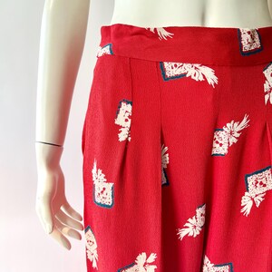 May include: Red high-waisted pants with a white floral and square pattern. The pants have pleats and a wide waistband. The fabric has a textured appearance. The pants are displayed on a mannequin.