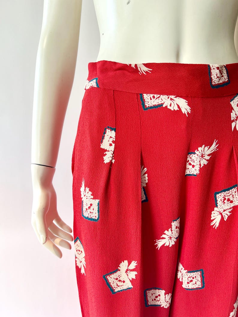May include: Red high-waisted pants with a white floral and square pattern. The pants have pleats and a wide waistband. The fabric has a textured appearance. The pants are displayed on a mannequin.