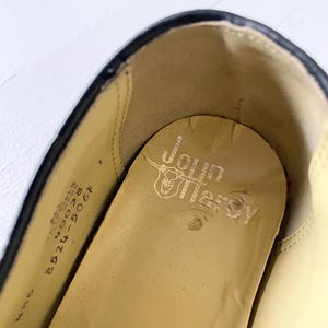May include: Close-up of the inside of a black leather shoe with a tan insole. The insole is stamped with the text "John  Herron" and a small bull's head logo. The shoe size is printed on the insole: "8 1/2  M  WOOD  8524-506F".