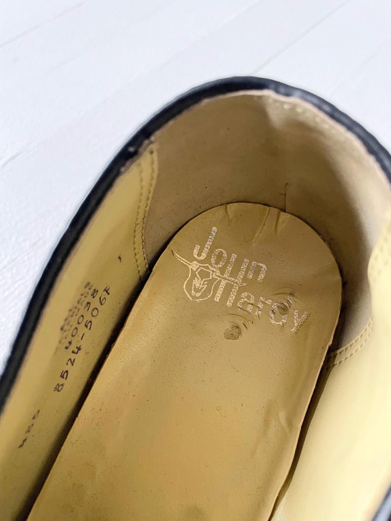 May include: Close-up of the inside of a black leather shoe with a tan insole. The insole is stamped with the text "John  Herron" and a small bull's head logo. The shoe size is printed on the insole: "8 1/2  M  WOOD  8524-506F".
