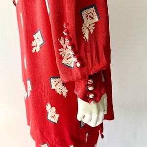 May include: A red dress with a white and blue patterned design. The dress features long sleeves with decorative buttons on the cuffs. The fabric has a textured appearance, and the design is repeated across the garment.