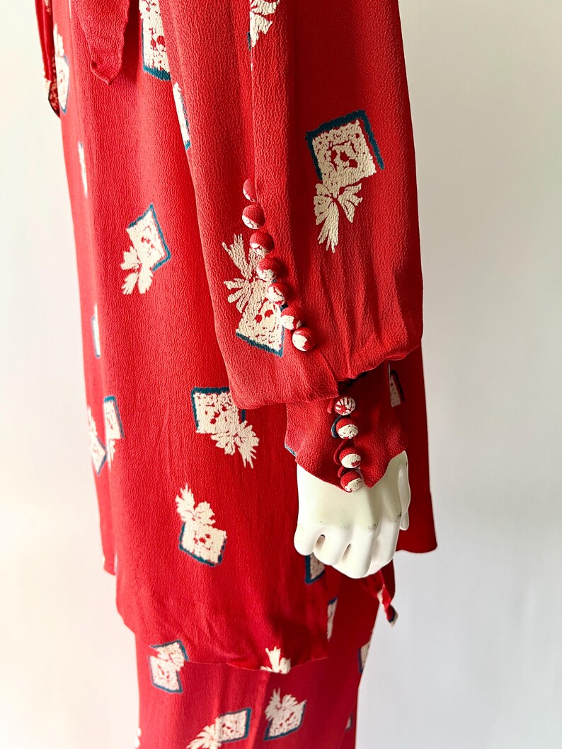May include: A red dress with a white and blue patterned design. The dress features long sleeves with decorative buttons on the cuffs. The fabric has a textured appearance, and the design is repeated across the garment.