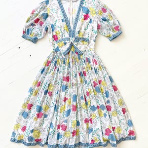 Puede incluir: Un vestido blanco con estampado floral en tonos azules, amarillos, rosas y verdes. El vestido tiene un escote en V, un lazo en la cintura y una falda fruncida. El vestido tiene mangas cortas abullonadas y un ribete azul y blanco.