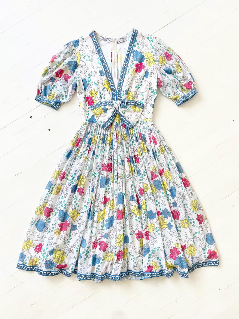 Puede incluir: Un vestido blanco con estampado floral en tonos azules, amarillos, rosas y verdes. El vestido tiene un escote en V, un lazo en la cintura y una falda fruncida. El vestido tiene mangas cortas abullonadas y un ribete azul y blanco.