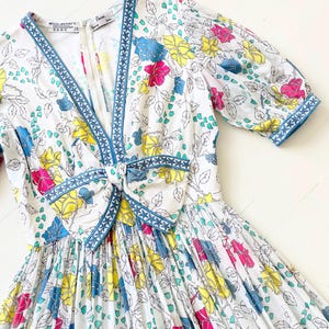 Puede incluir: Un vestido blanco con estampado floral en tonos de rosa, amarillo, azul y verde. El vestido tiene un escote en V y un ribete azul alrededor del escote y las mangas. El vestido tiene un lazo en la cintura.