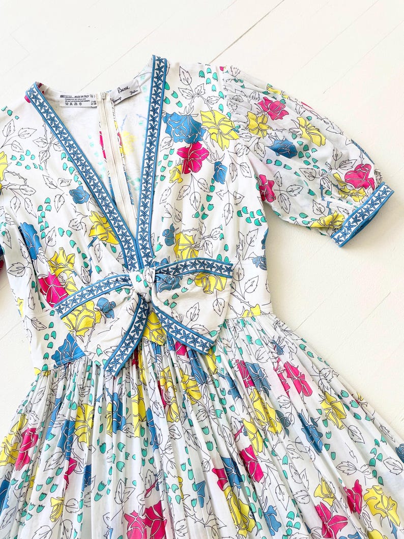 Puede incluir: Un vestido blanco con estampado floral en tonos de rosa, amarillo, azul y verde. El vestido tiene un escote en V y un ribete azul alrededor del escote y las mangas. El vestido tiene un lazo en la cintura.