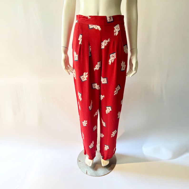 May include: Red high-waisted pants with a white and blue pattern. The pants have a pleated front and a wide leg. The fabric appears to be lightweight and suitable for warmer weather. The pattern includes floral and geometric shapes.