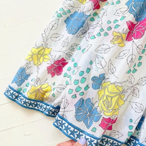 Puede incluir: Tela de algod&oacute;n blanca con estampado floral con rosas azules, amarillas y rosas. La tela tiene un ribete azul con un patr&oacute;n de estrellas blancas.