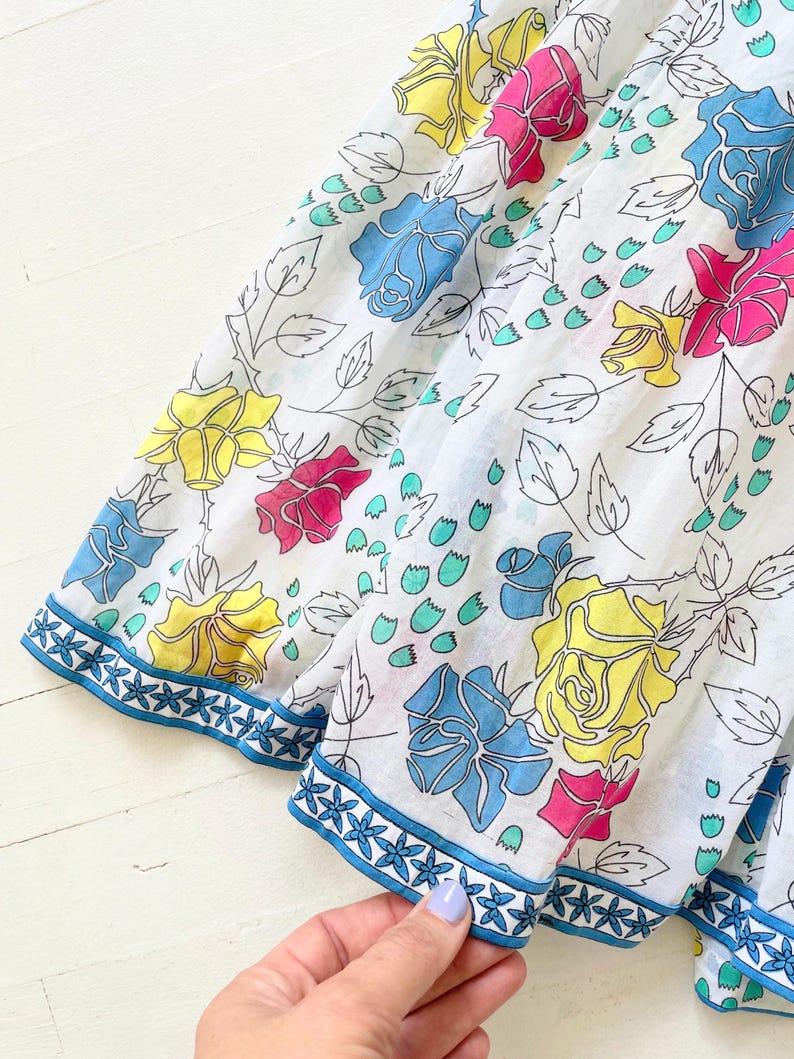 Puede incluir: Tela de algod&oacute;n blanca con estampado floral con rosas azules, amarillas y rosas. La tela tiene un ribete azul con un patr&oacute;n de estrellas blancas.
