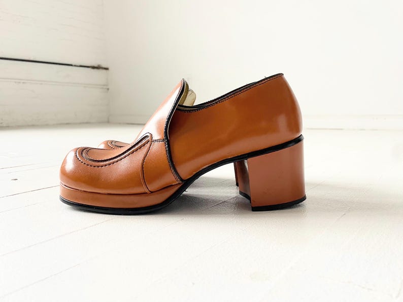 May include: Brown leather platform shoes with a chunky heel. The shoes have a pointed toe and a decorative seam detail.