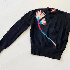 May include: Black wrap-style jumper with a floral design in white, red, and blue. The jumper has long sleeves and a V-neck. The floral design is on the left side.