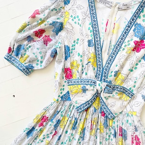 Puede incluir: Un vestido blanco con estampado floral con flores rosas, amarillas, azules y verdes. El vestido tiene un escote en V y un ribete azul con un patr&oacute;n geom&eacute;trico blanco. El vestido tiene un lazo en la cintura y una cremallera en la espalda. El vestido est&aacute; hecho en Italia por la marca "Esca".