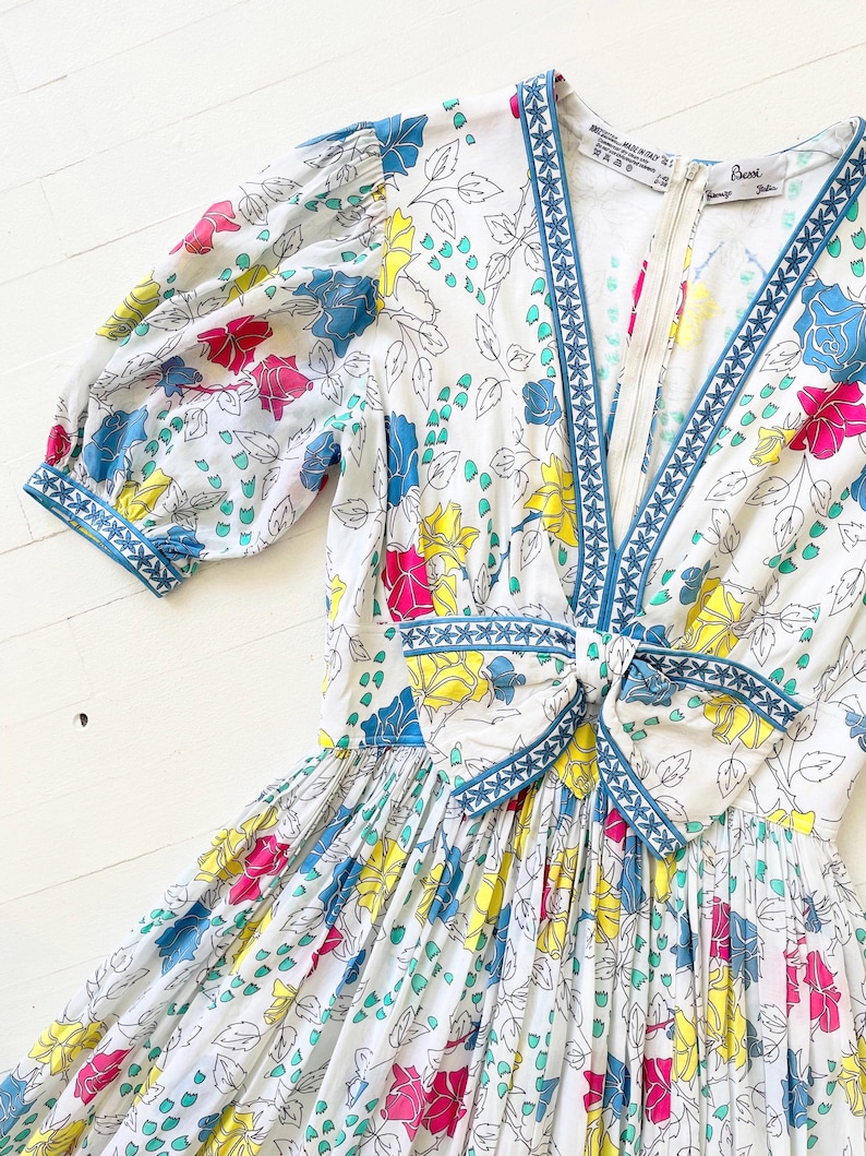 Puede incluir: Un vestido blanco con estampado floral con flores rosas, amarillas, azules y verdes. El vestido tiene un escote en V y un ribete azul con un patr&oacute;n geom&eacute;trico blanco. El vestido tiene un lazo en la cintura y una cremallera en la espalda. El vestido est&aacute; hecho en Italia por la marca "Esca".