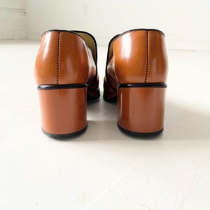 May include: A pair of brown leather heeled loafers with a black trim. The shoes have a block heel and a pointed toe.