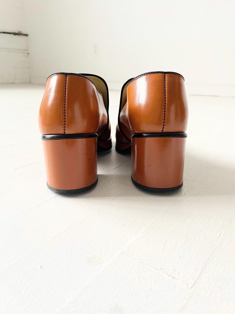 May include: A pair of brown leather heeled loafers with a black trim. The shoes have a block heel and a pointed toe.