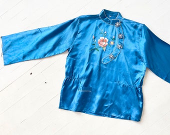 Puede incluir: Una bata de seda azul con un diseño de bordado floral y botones decorativos. La bata tiene un cuello mandarín y mangas largas.