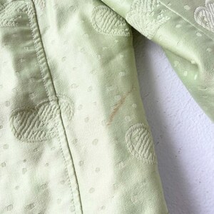 Puede incluir: Primer plano de una chaqueta verde claro con una superficie texturizada. La chaqueta presenta dise&ntilde;os circulares y florales bordados, junto con peque&ntilde;os patrones de puntos. Una mancha marr&oacute;n es visible en la tela.