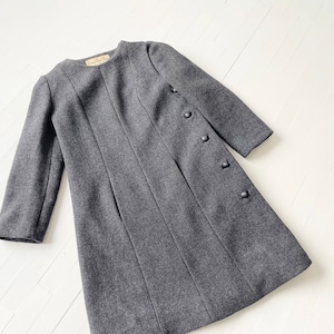 May include: A gray wool coat with a round neckline and a row of buttons down the side. The coat has a simple, classic design.