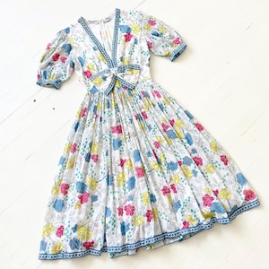 Puede incluir: Un vestido midi blanco con estampado floral en tonos rosa, azul y amarillo. El vestido tiene un escote en V, mangas abullonadas cortas y un lazo en la cintura.