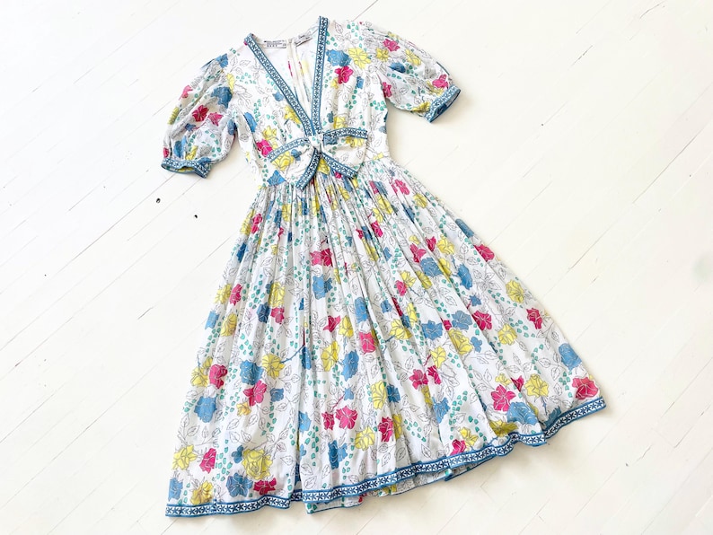Puede incluir: Un vestido midi blanco con estampado floral en tonos rosa, azul y amarillo. El vestido tiene un escote en V, mangas abullonadas cortas y un lazo en la cintura.