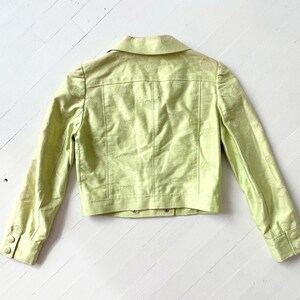 Puede incluir: Chaqueta verde claro con cuello y mangas largas. La chaqueta presenta un sutil estampado floral y botones en los pu&ntilde;os y en la parte delantera. Se muestra sobre una superficie de madera blanca.