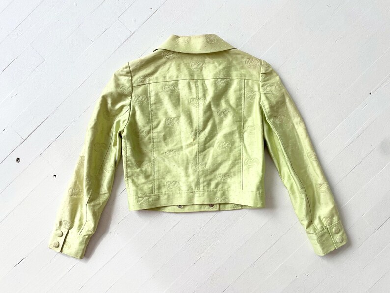 Puede incluir: Chaqueta verde claro con cuello y mangas largas. La chaqueta presenta un sutil estampado floral y botones en los pu&ntilde;os y en la parte delantera. Se muestra sobre una superficie de madera blanca.