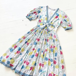 Puede incluir: Un vestido blanco con estampado floral en tonos de rosa, amarillo, azul y verde. El vestido tiene un escote en V, mangas cortas abullonadas y un lazo en la cintura.