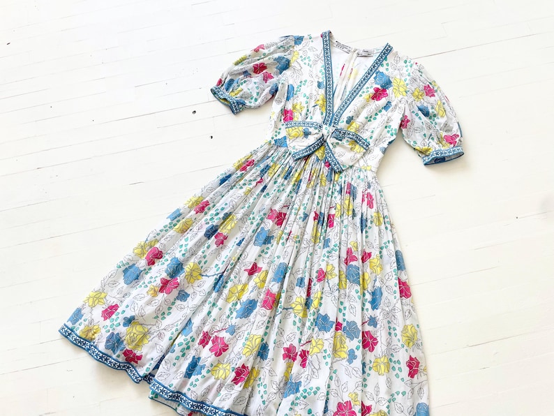 Puede incluir: Un vestido blanco con estampado floral en tonos de rosa, amarillo, azul y verde. El vestido tiene un escote en V, mangas cortas abullonadas y un lazo en la cintura.