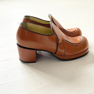 May include: A pair of brown leather platform shoes with a chunky heel. The shoes have a simple design with a single seam across the toe.
