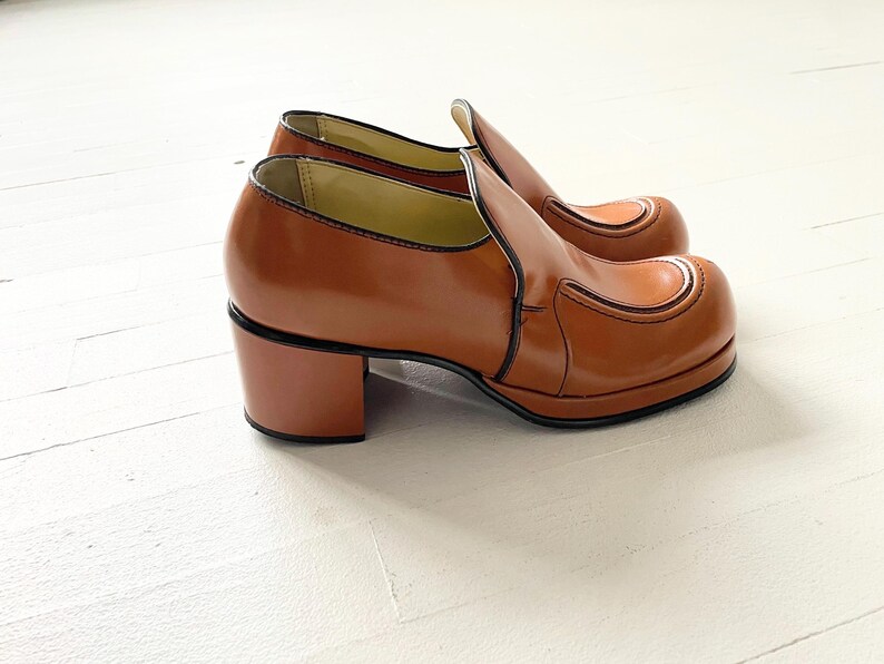 May include: A pair of brown leather platform shoes with a chunky heel. The shoes have a simple design with a single seam across the toe.