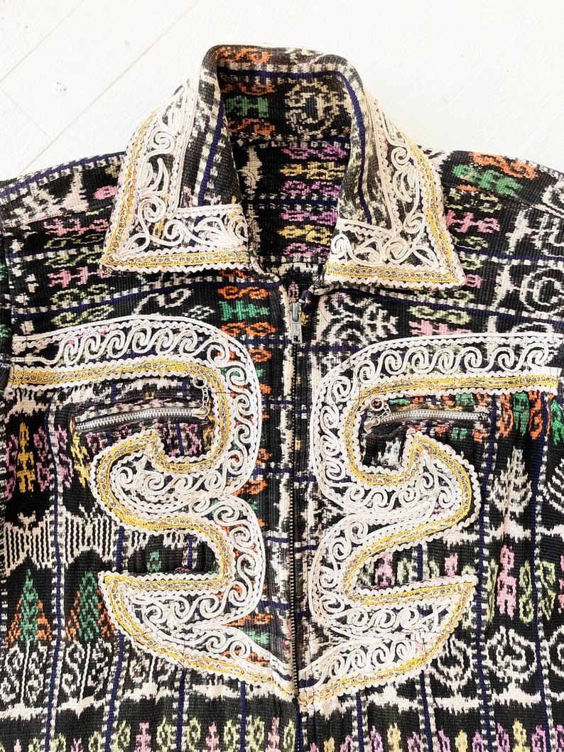 Puede incluir: Una chaqueta con estampado negro y blanco con bordados dorados y blancos. La chaqueta tiene cierre de cremallera y cuello alto.