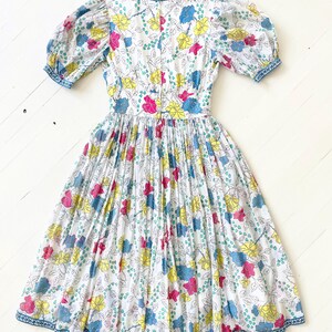 Puede incluir: Un vestido blanco con estampado floral en tonos rosa, amarillo, azul y verde. El vestido tiene una falda fruncida y mangas abullonadas con un ribete azul.