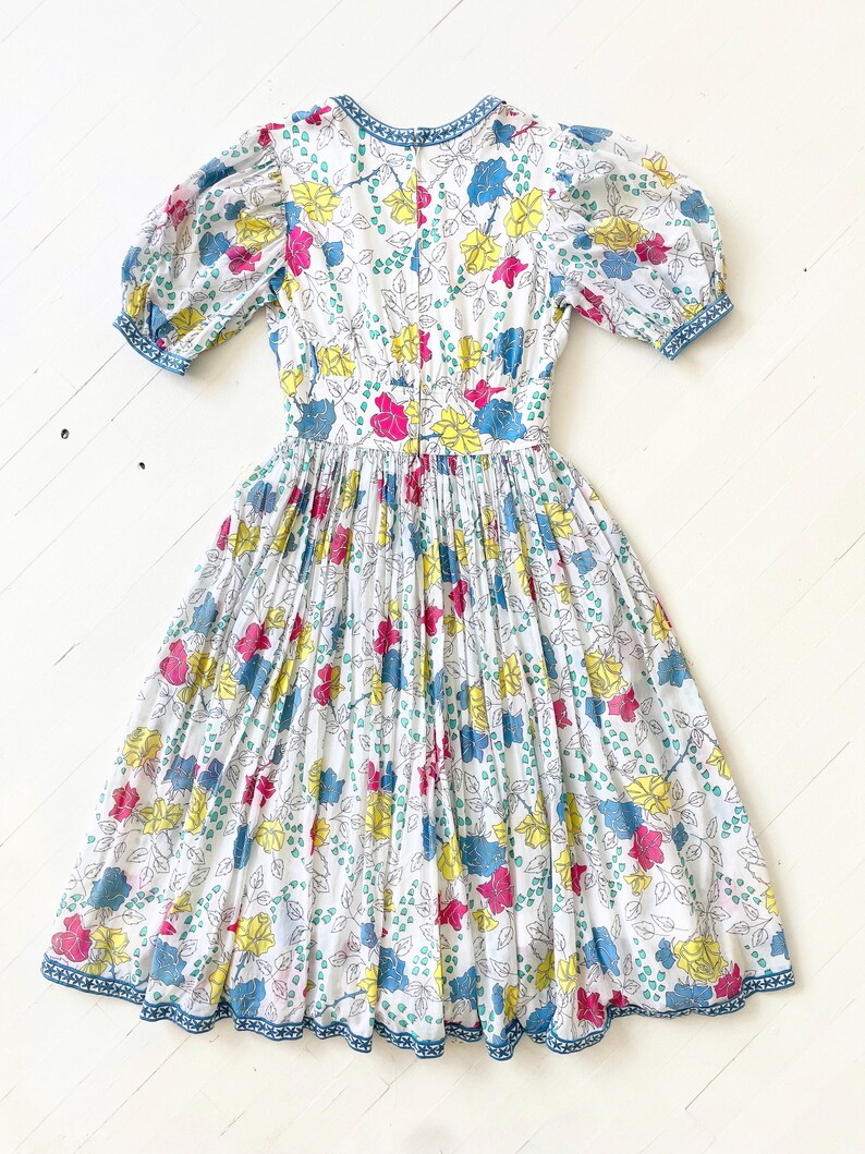 Puede incluir: Un vestido blanco con estampado floral en tonos rosa, amarillo, azul y verde. El vestido tiene una falda fruncida y mangas abullonadas con un ribete azul.