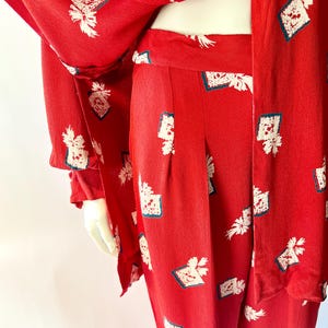 May include: A red two-piece outfit featuring a jacket and pants. The fabric is patterned with white floral and geometric designs with teal accents. The jacket has long sleeves and buttons. The pants have a high waist and a straight leg.